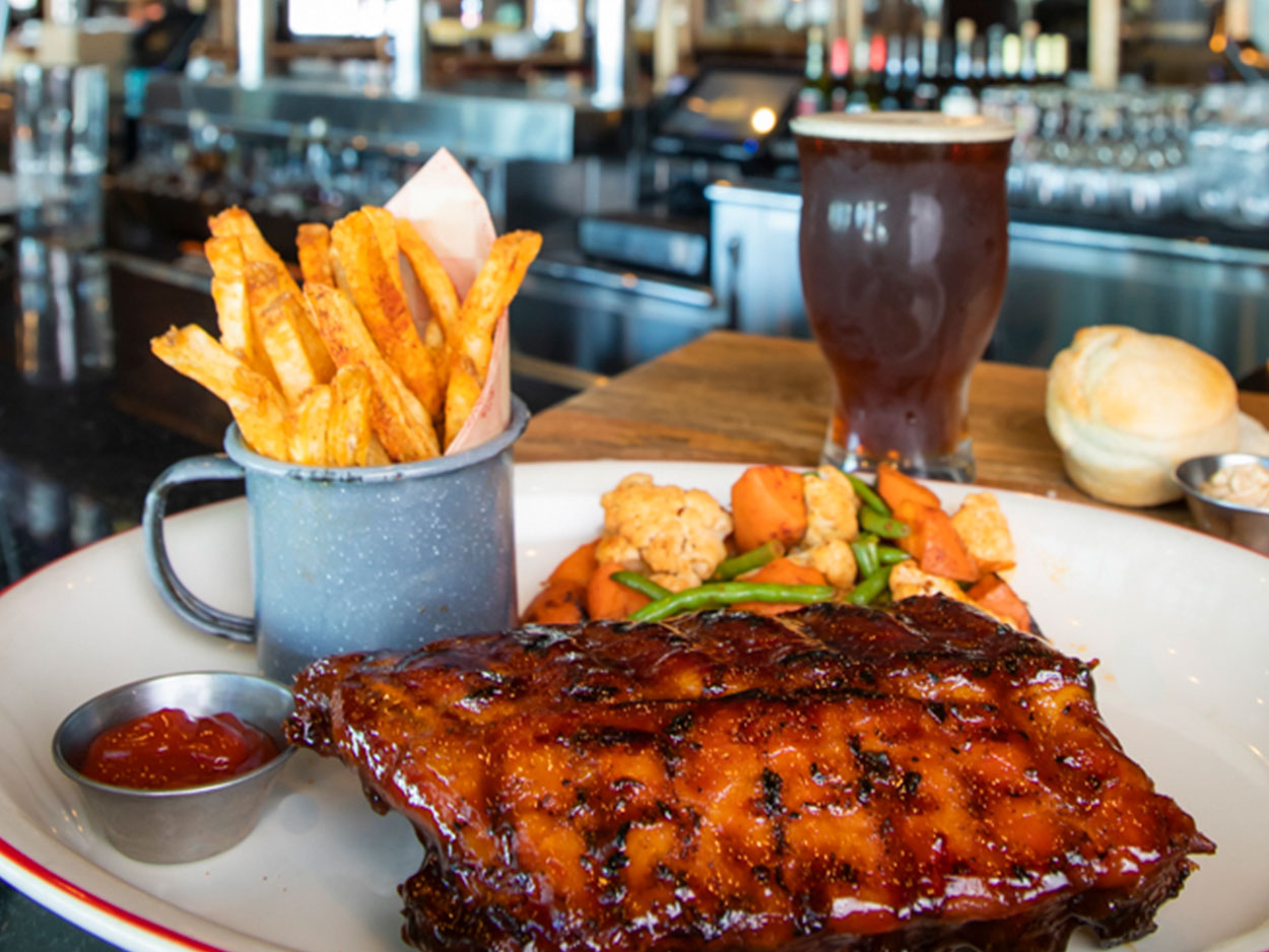 Half rack of ribs with an amber-colored pint of beer, mixed vegetables, and a side of fries at the bar top of the Lucille's Flying Pig Lounge.